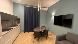 A modern kitchen with a dining area, a flat-screen TV mounted on the wall, and a sofa in the living area.