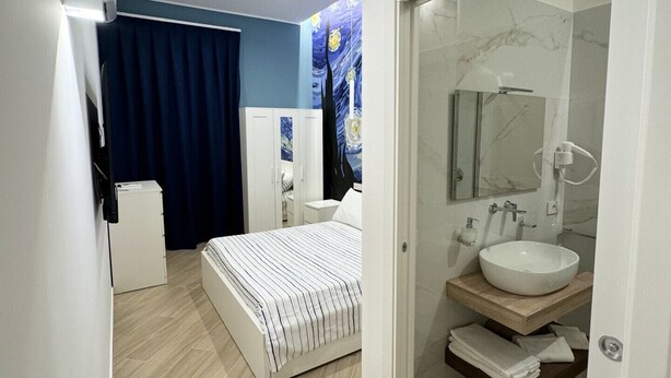 A modern bedroom with a bed, a small white cabinet, a mirror, and a bathroom with a sink and marble tiles.