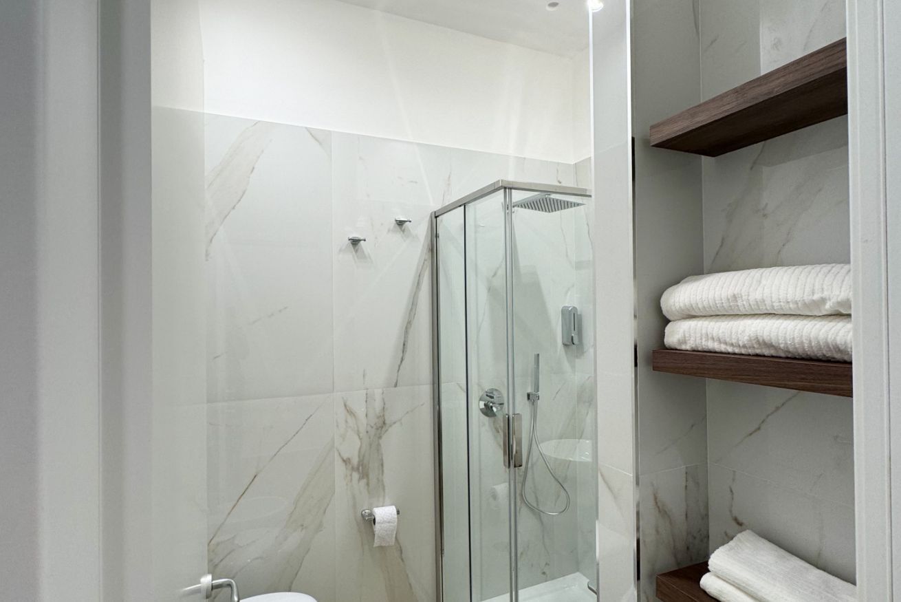 A modern bathroom with a glass-enclosed shower, marble walls, and a wooden shelf with towels.