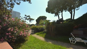 A pathway leading through a garden with various plants and trees, a white chair, and a building in the background. - Comfy Beachside Vacation Home With Patio and Garden (Lignano Sabbiadoro)