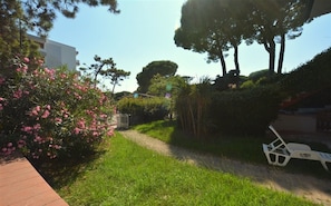 Un sentier qui traverse un jardin où l’on trouve différentes plantes et arbres, une chaise blanche et, au loin, un bâtiment.