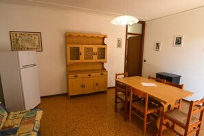 A compact room with a wooden dining table and chairs, a white refrigerator, and a cabinet with patterned doors. - Comfy Beachside Vacation Home With Patio and Garden (Lignano Sabbiadoro)