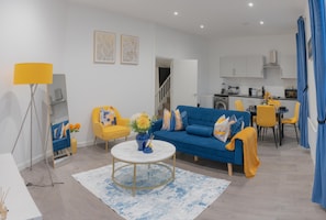 A modern living room with a blue sofa, yellow chairs, a round coffee table, and a dining area with a kitchen in the background. - Cosy City Apartment in Leicester (Leicester)