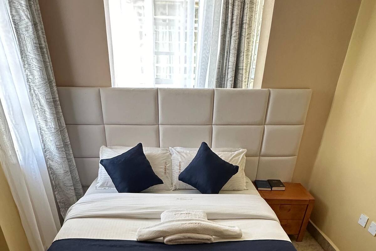 Un lit bien fait, avec une tête de lit, deux oreillers, une serviette pliée et une table de chevet sur laquelle sont posés des livres.