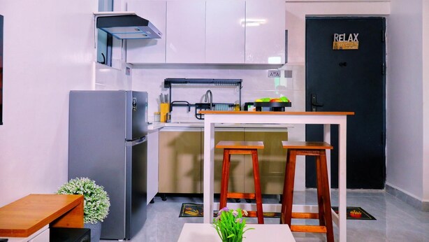 A compact kitchen with white cabinets, a black door labeled "RELAX," a stainless steel refrigerator, and a small dining area with stools. - Comfort home! made life simple, Beautiful views and desirable lifestyle. (Nairobi)