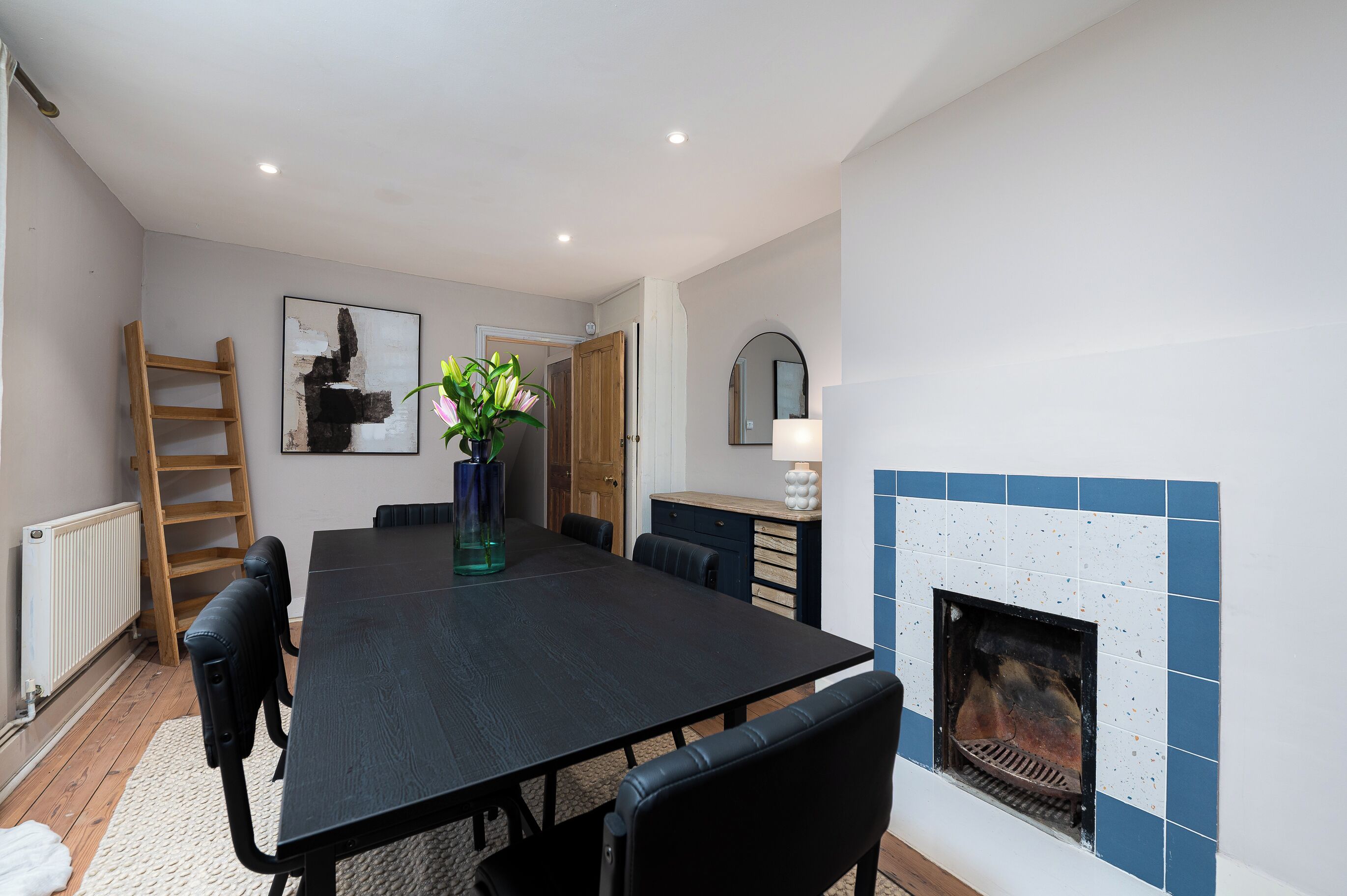 A dining room with a black table, chairs, a vase with flowers, a wooden ladder, a painting, a mirror, and a tiled fireplace.