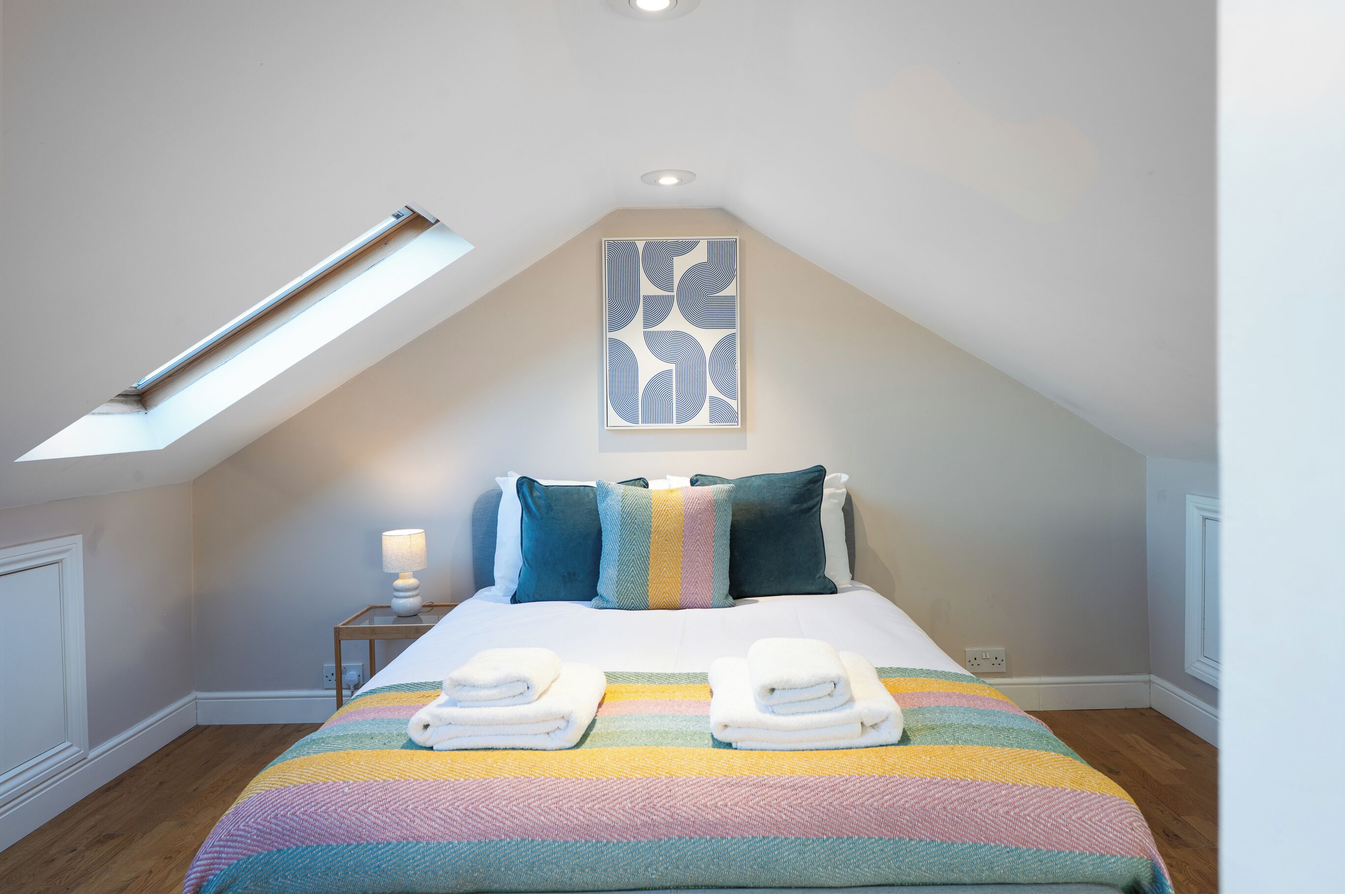 A neatly made bed with two towels, a bedside lamp, and a ceiling light in a room with a sloped ceiling and a skylight.