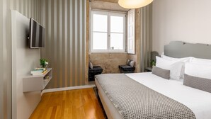 Una habitación de hotel moderna con una cama grande, un televisor montado en la pared, un pequeño rincón de descanso con una mesa y sillas, y un ventanal que deja entrar luz natural.
