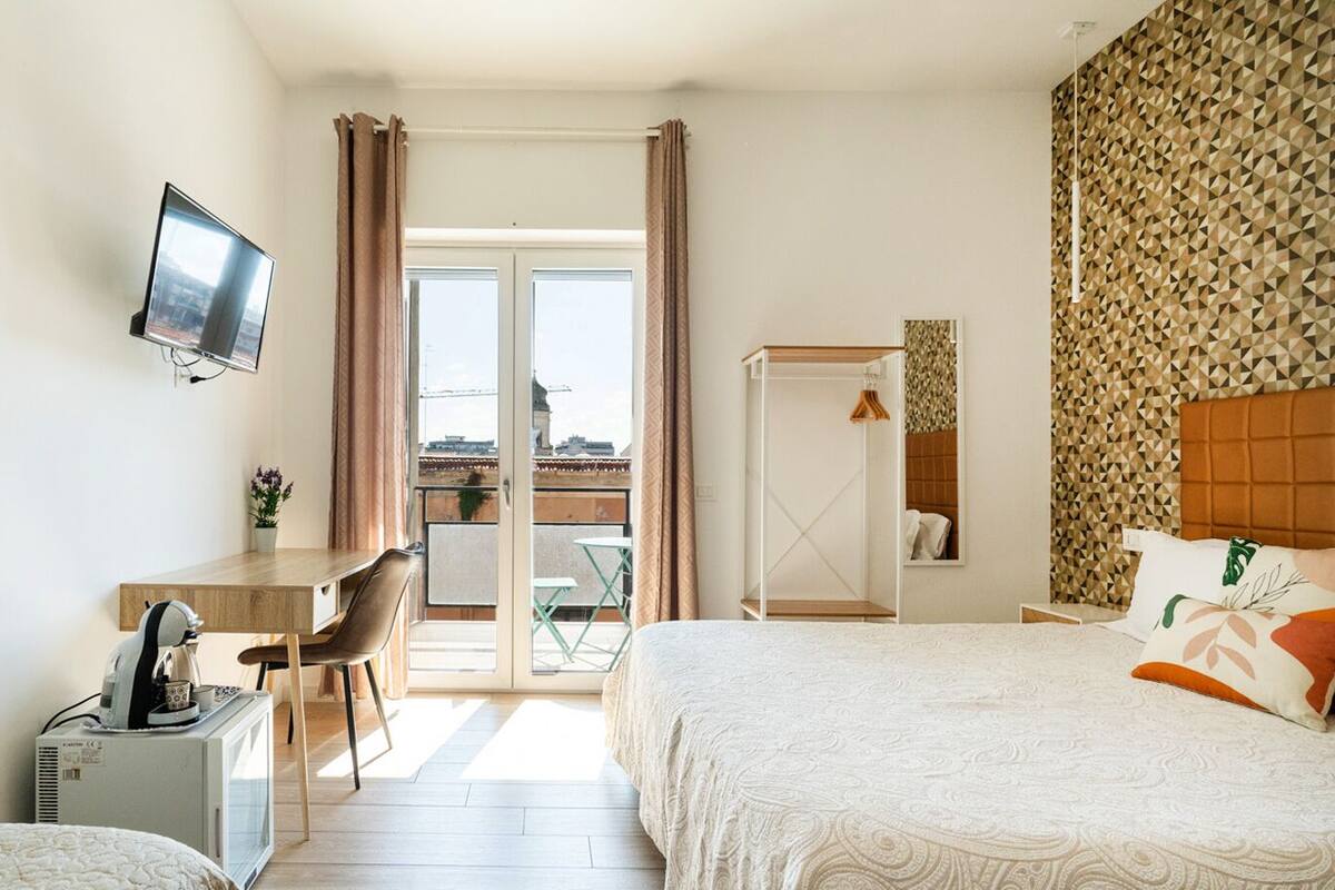 Habitación de hotel con cama, escritorio con silla, televisión y vista a la ciudad a través de un ventanal grande.