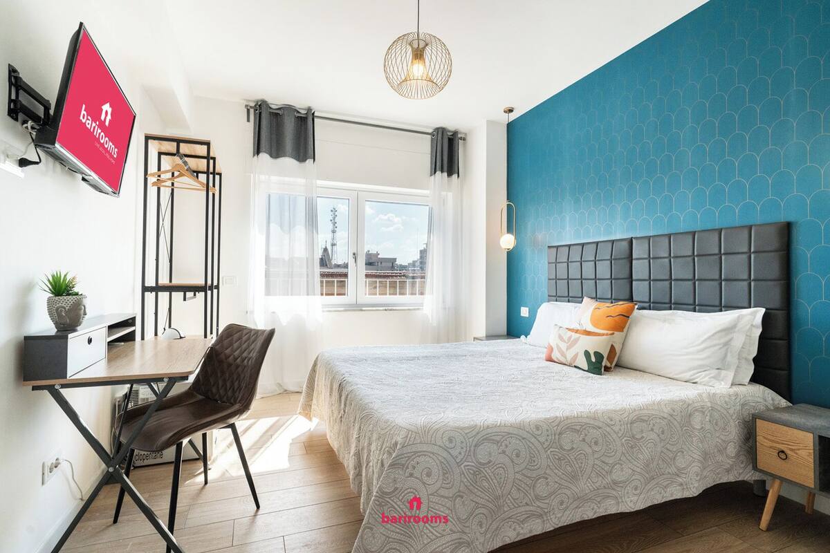 Habitación de hotel moderna con cama, mesita de noche y escritorio con silla. Hay un televisor montado en la pared, una ventana con cortinas y una pared decorada en azul.