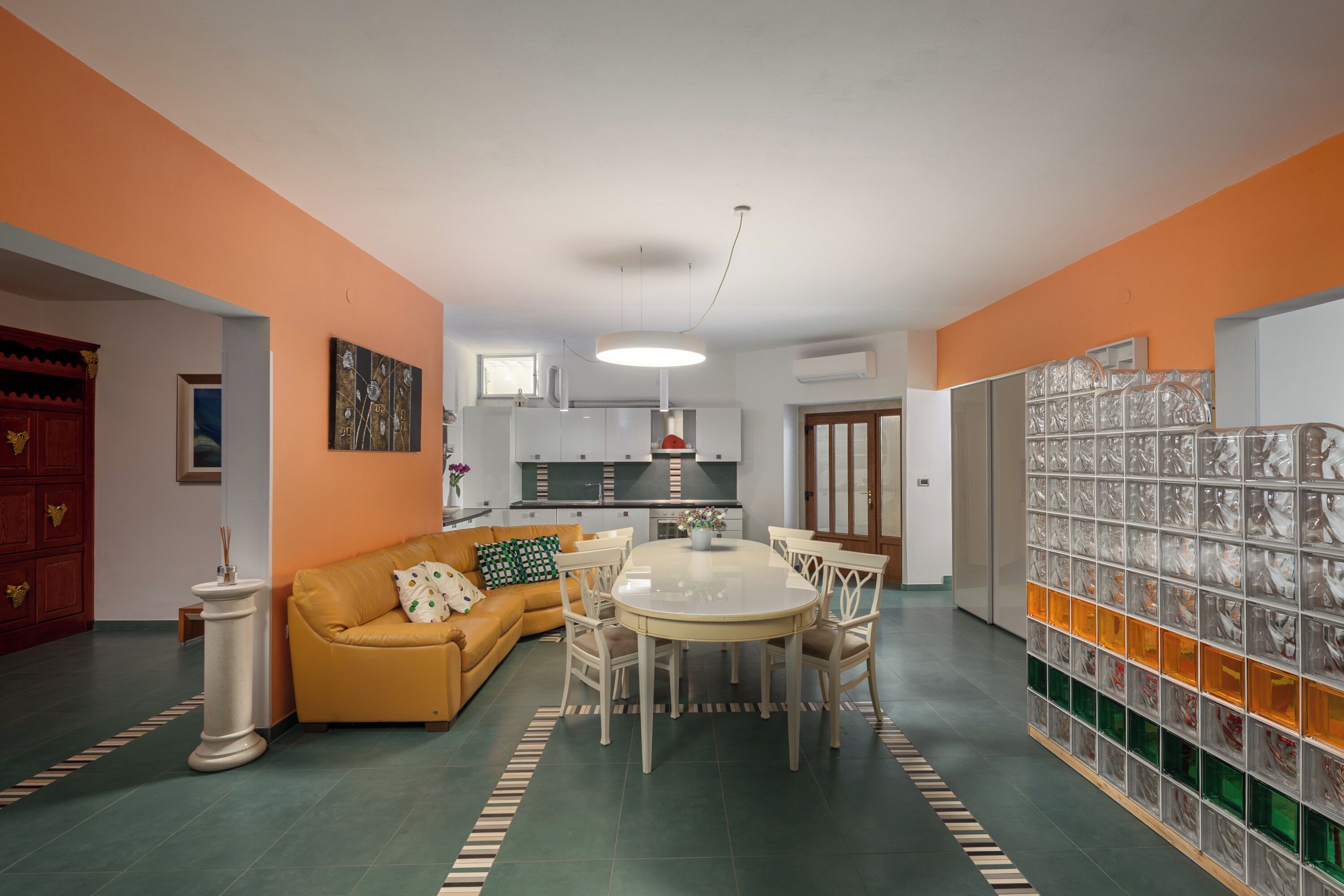 A living room with a sofa, dining table, and chairs, a kitchen in the background, and a decorative glass wall.