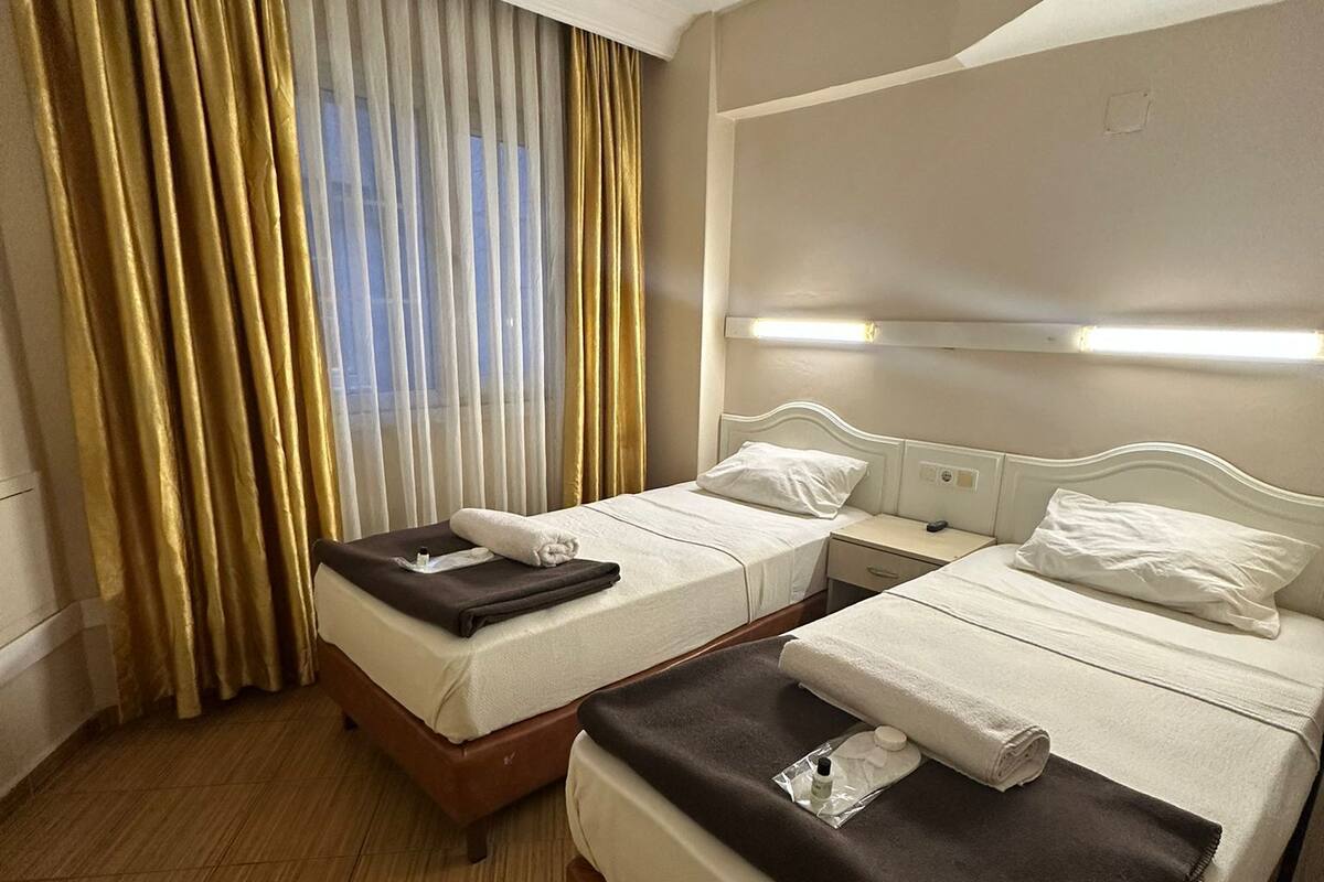 A hotel room with two beds, a large window with curtains, a bedside table, and a headboard with a light fixture.