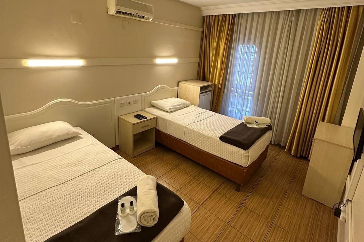 A hotel room with two beds, a bedside table with a lamp, an air conditioning unit, a flat-screen TV, and a window with curtains.