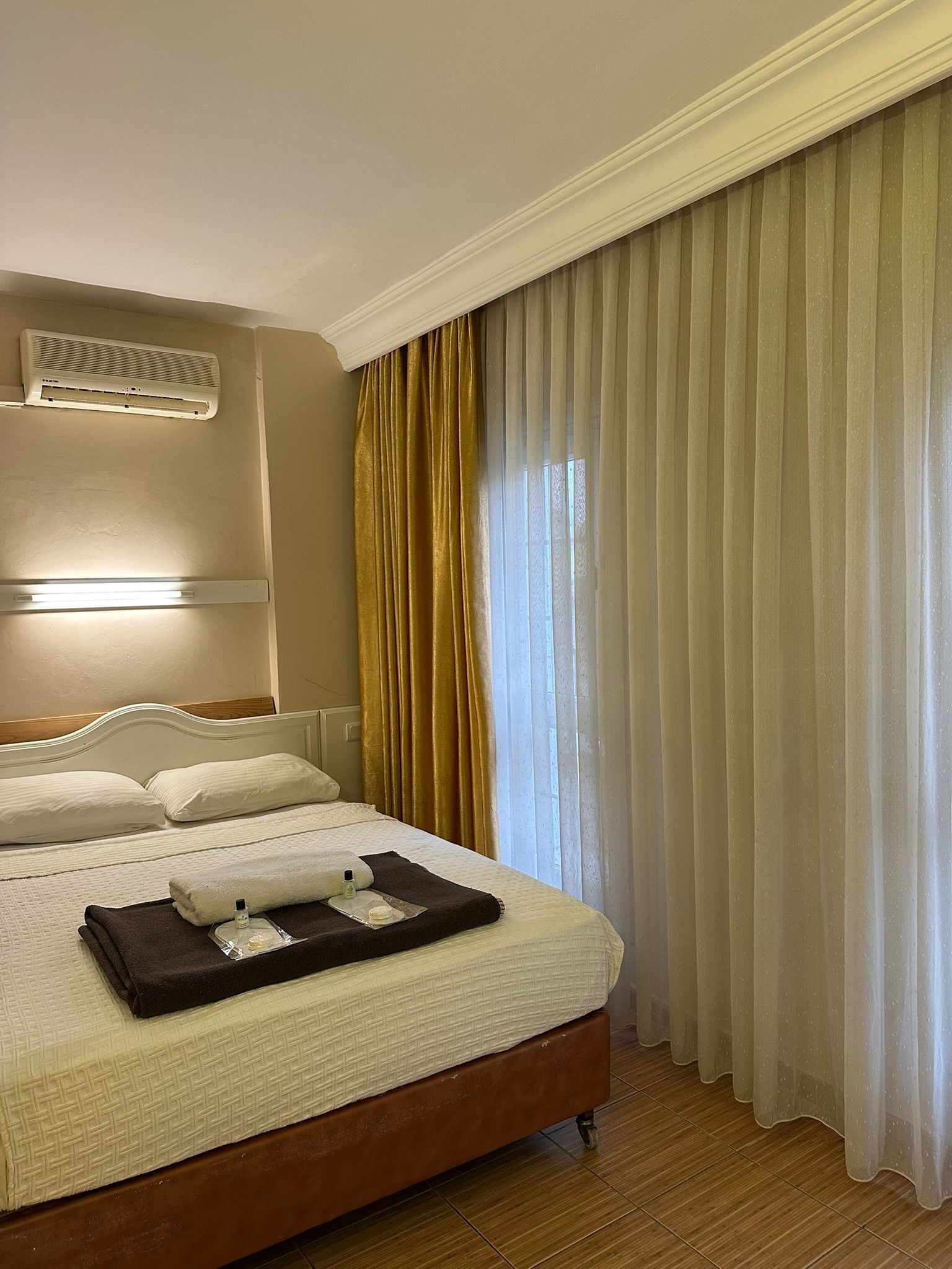 Ein Hotelzimmer mit Bett, Nachttisch, Lampe, Fenster mit Vorhängen und Klimaanlage.