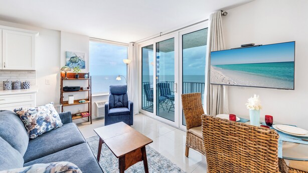 A modern living room with a sofa, a coffee table, and a dining area with a table and chairs. There is a large window with a view of the ocean.