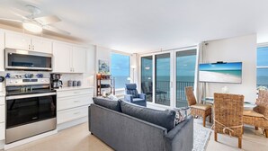 A modern kitchen with white cabinets, a built-in oven, and a microwave. There is a dining area with wicker chairs and a table, and a large window offering a view of the ocean.