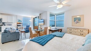 A modern hotel room with a large bed, a desk, a dining area, and a view of the ocean.