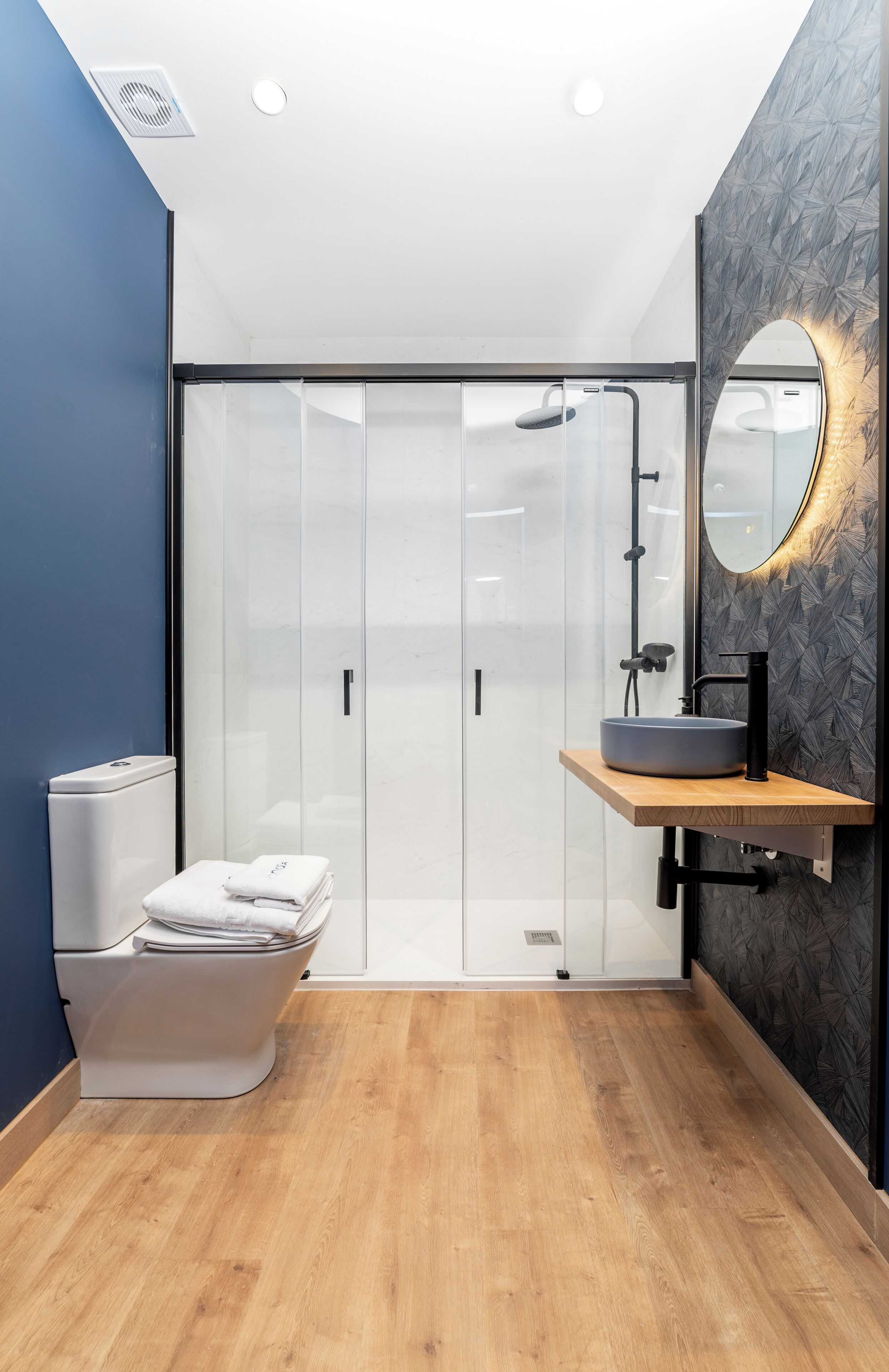 Ein modernes Badezimmer mit Glasabtrennung, Toilette in Weiß, Waschbecken mit schwarzem Wasserhahn und Holzkommode.