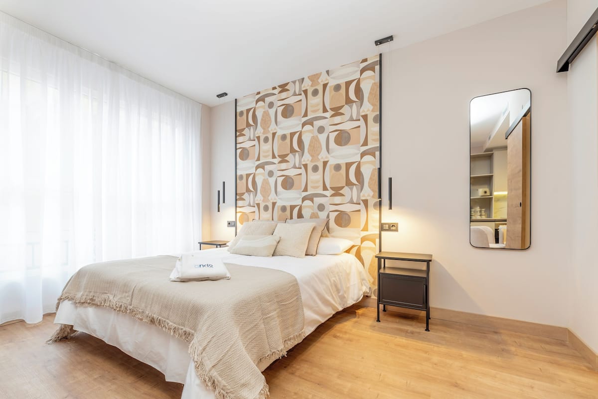 Une chambre moderne avec un grand lit, une table de chevet, un miroir fixé au mur et un mur orné d’un motif géométrique.