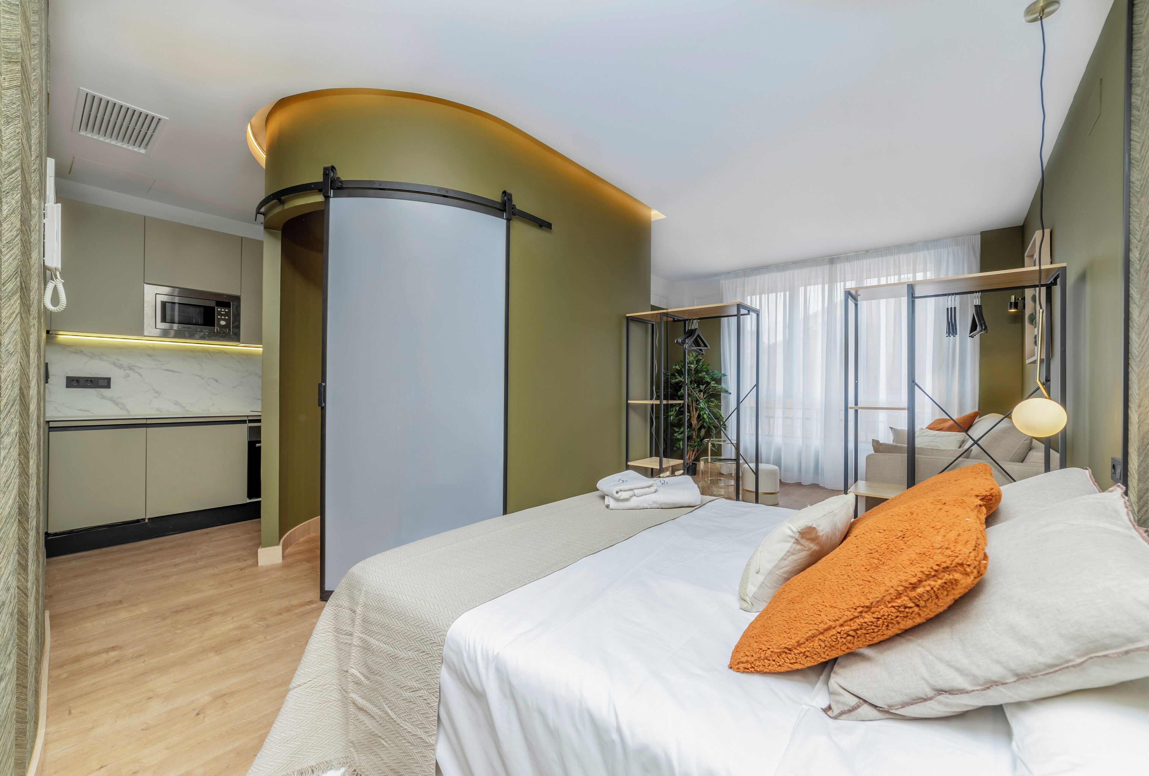 Quarto de hotel moderno com cama, uma kitchenette pequena, banheiro com chuveiro de vidro e vista para a cidade.