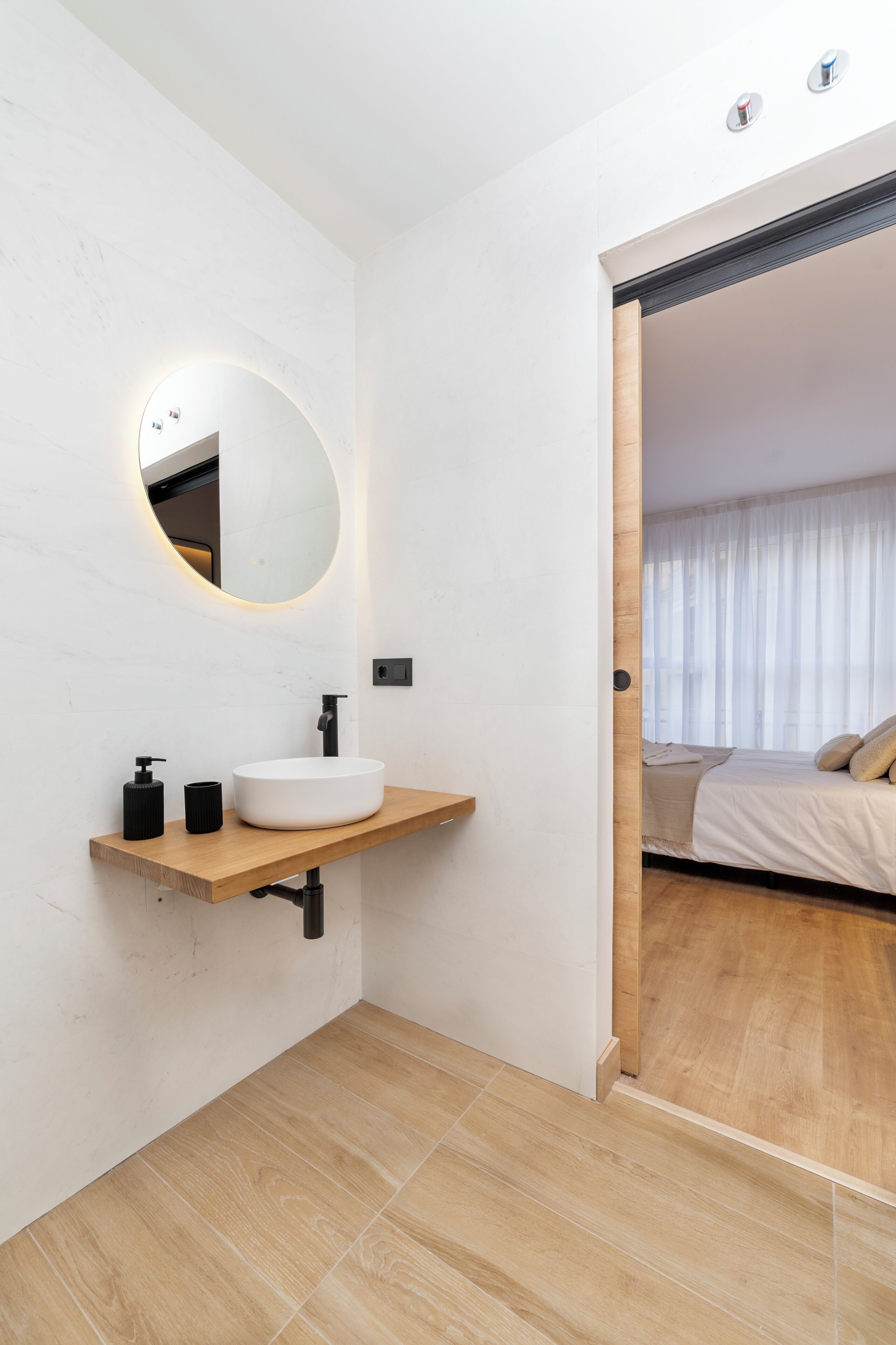 A modern bathroom with a white sink, a round mirror, and a wooden shelf.
