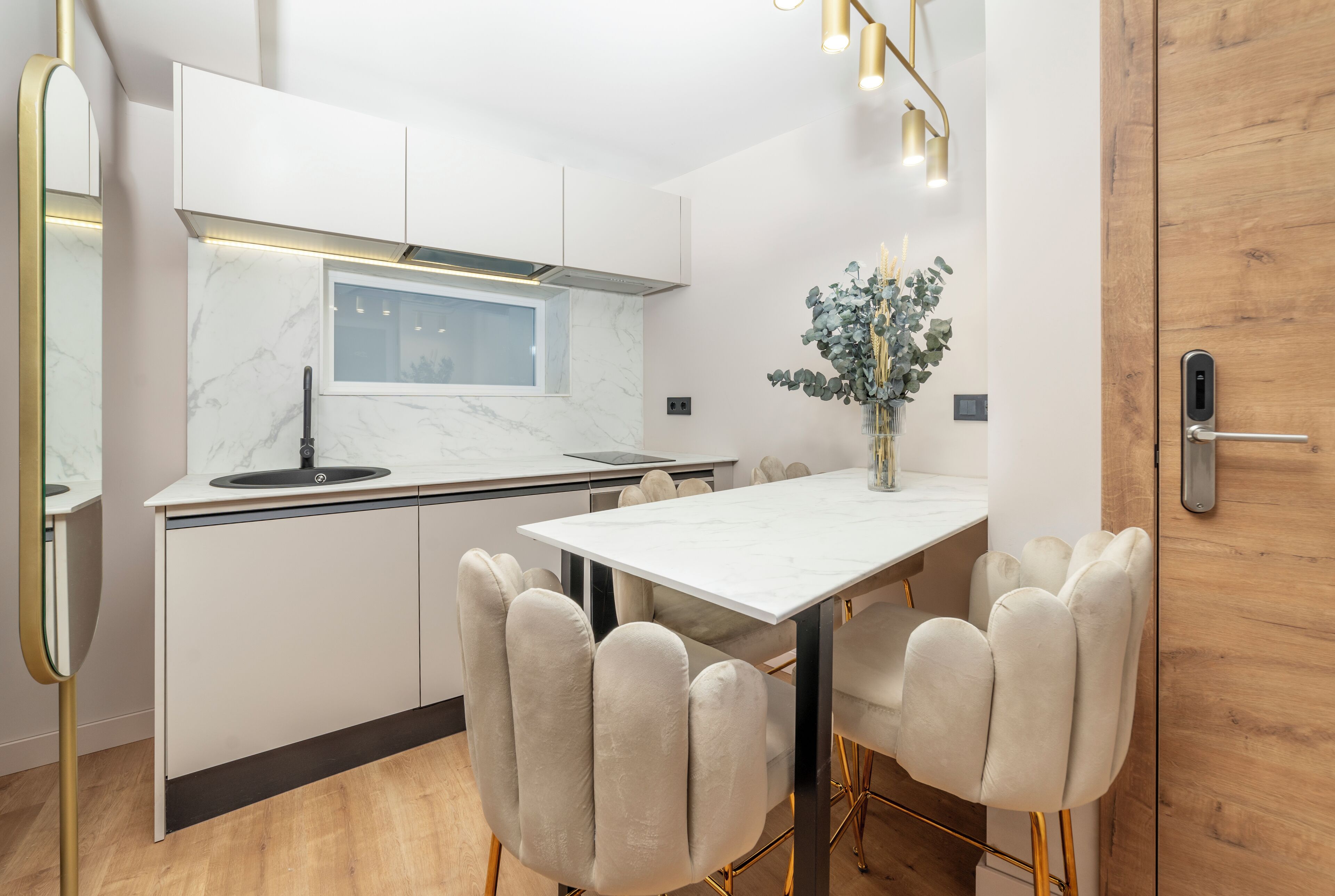Une cuisine moderne avec des meubles blancs, un pare-feu en marbre, une fenêtre intégrée et un coin repas comprenant une table blanche et des chaises rembourrées.