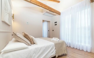 Un dormitorio con cama, almohadas, una mesita de noche, una ventana con cortinas y una viga de techo de madera.