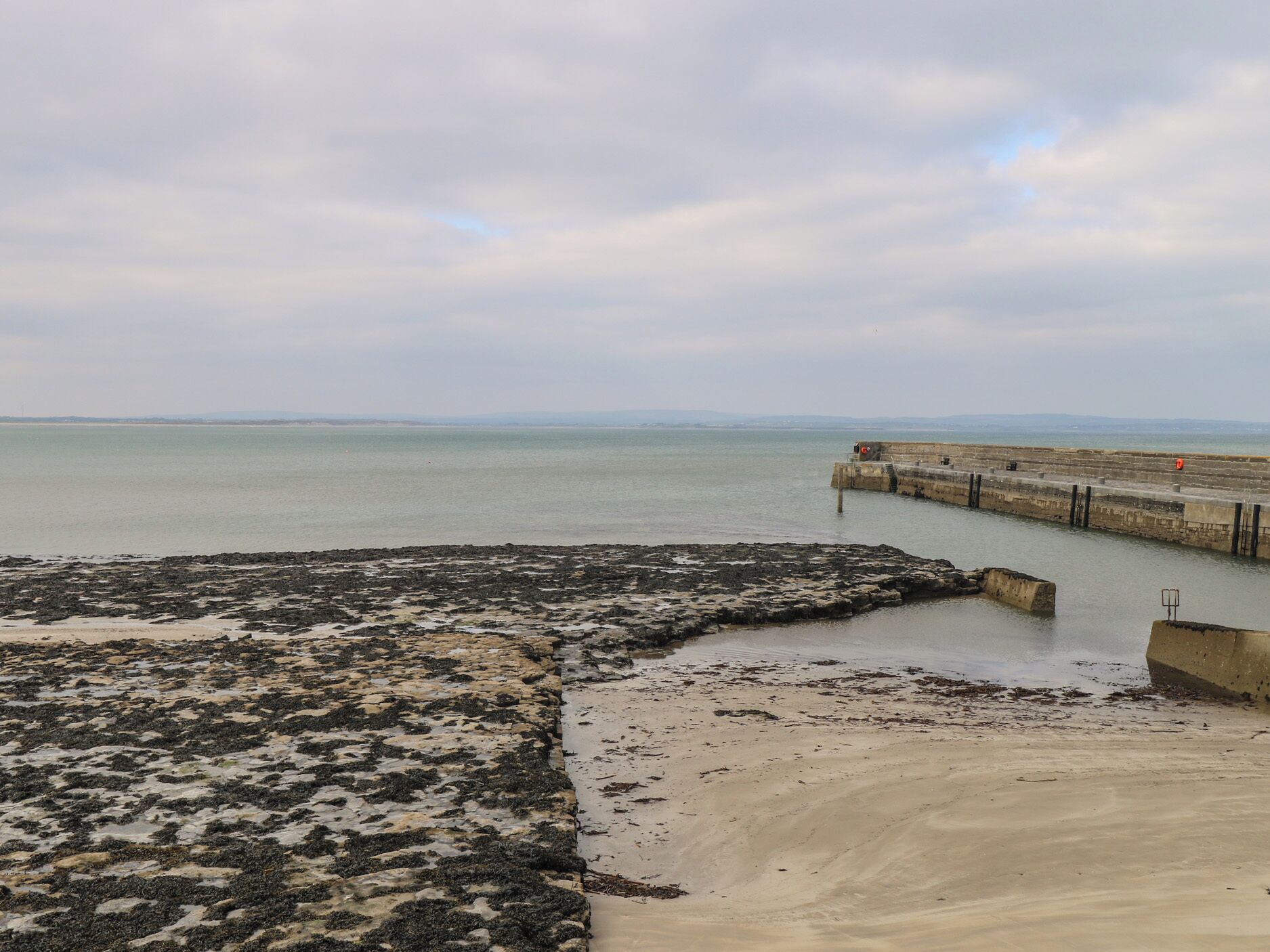 A coastal view with a concrete pier extending into calm waters, a rocky beach area, and a distant shoreline.