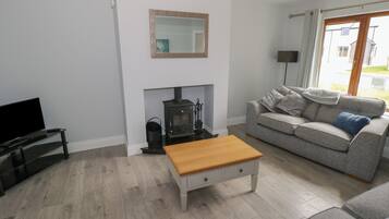 Una sala de estar con un sofá gris, una mesa de centro de madera, un televisor sobre un mueble, una chimenea y una ventana con cortinas.