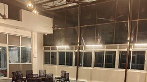 A ping pong table with two paddles, surrounded by chairs, in a room with large windows and ceiling fans. - Glow Inn HomeStay (Varanasi)