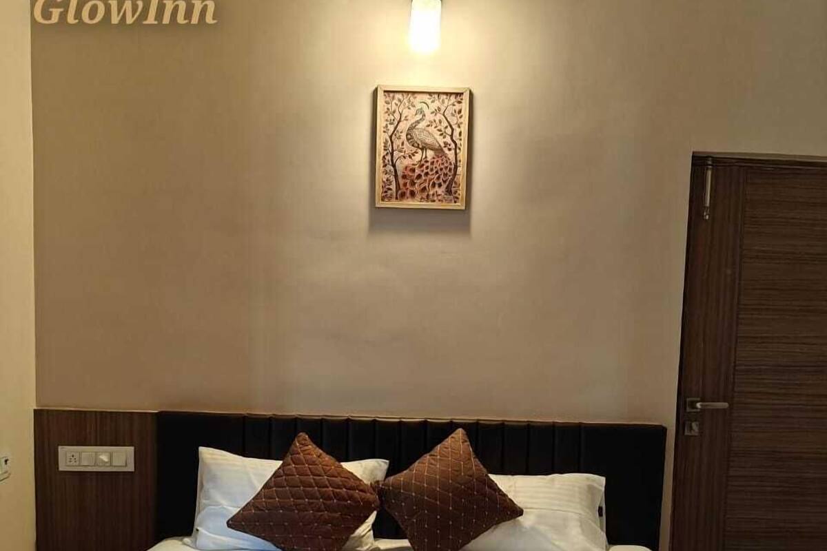 Una habitación de hotel con una cama, dos almohadas, una lámpara fijada a la pared, un cuadro enmarcado y una puerta.