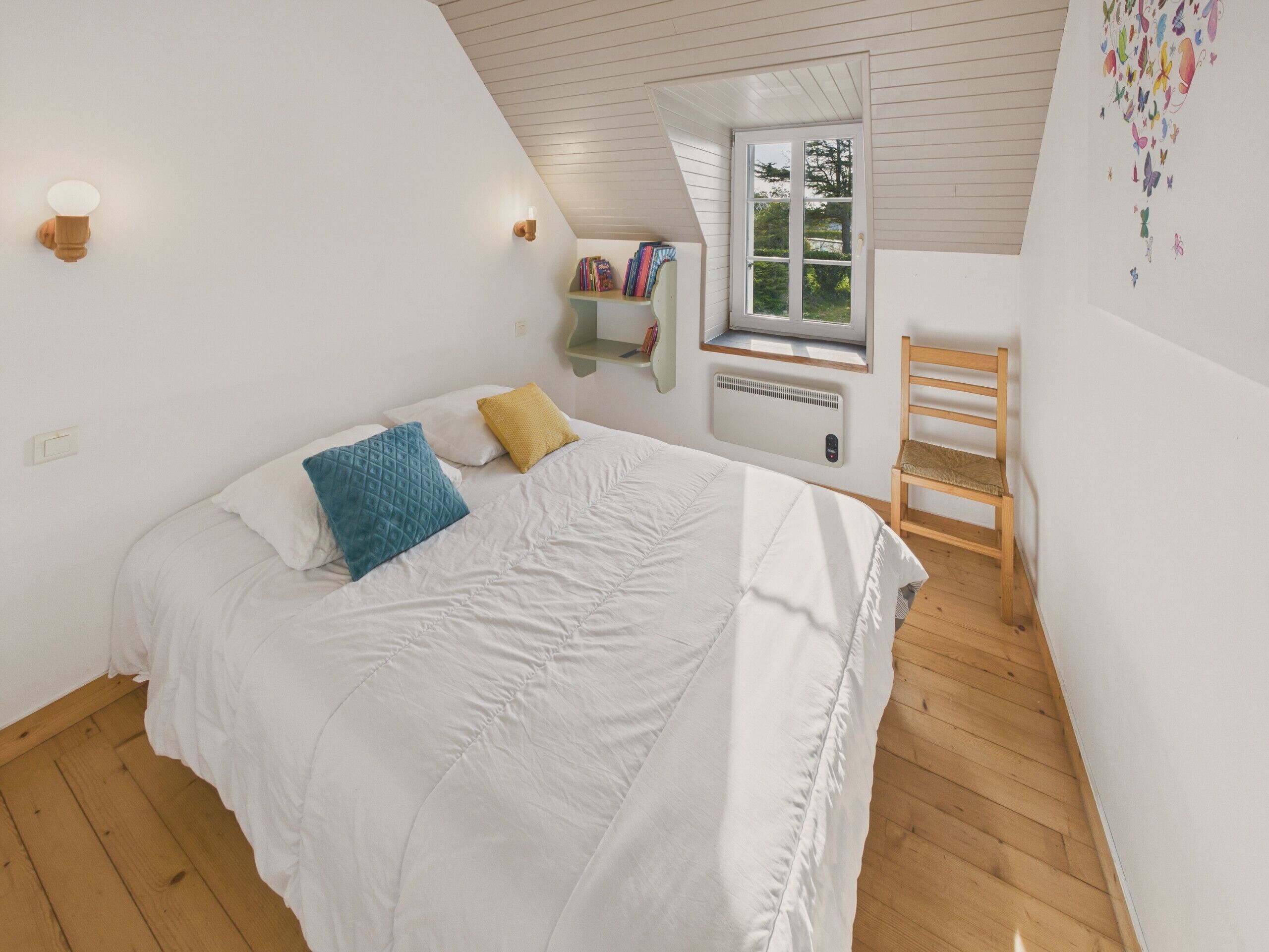 A bedroom with a sloped ceiling, a window with a view of trees, a bed with white bedding and two pillows, a wooden chair, and a wall-mounted shelf with books.