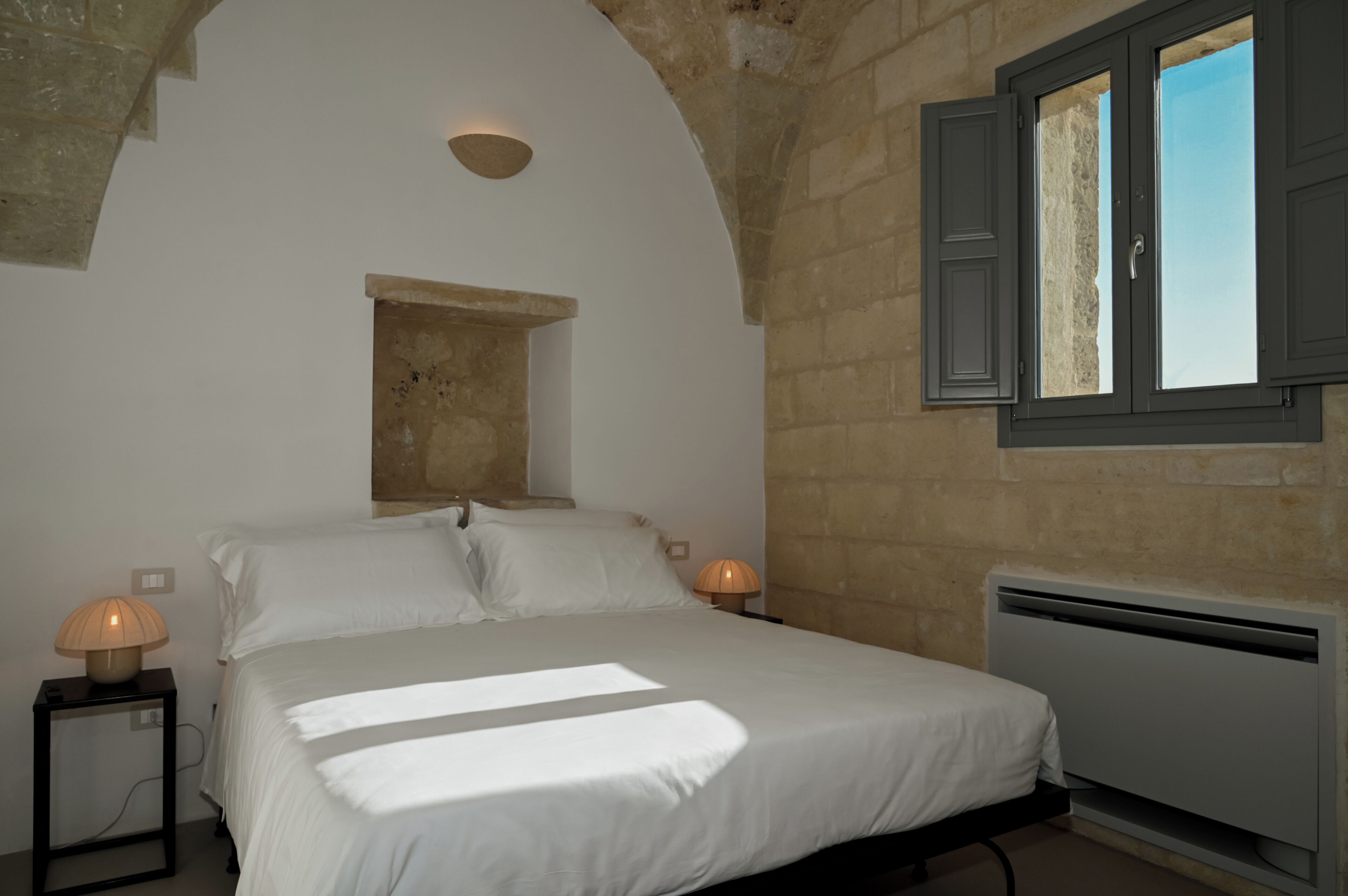 A bedroom with a bed, two bedside lamps, a small window with shutters, and a stone wall.
