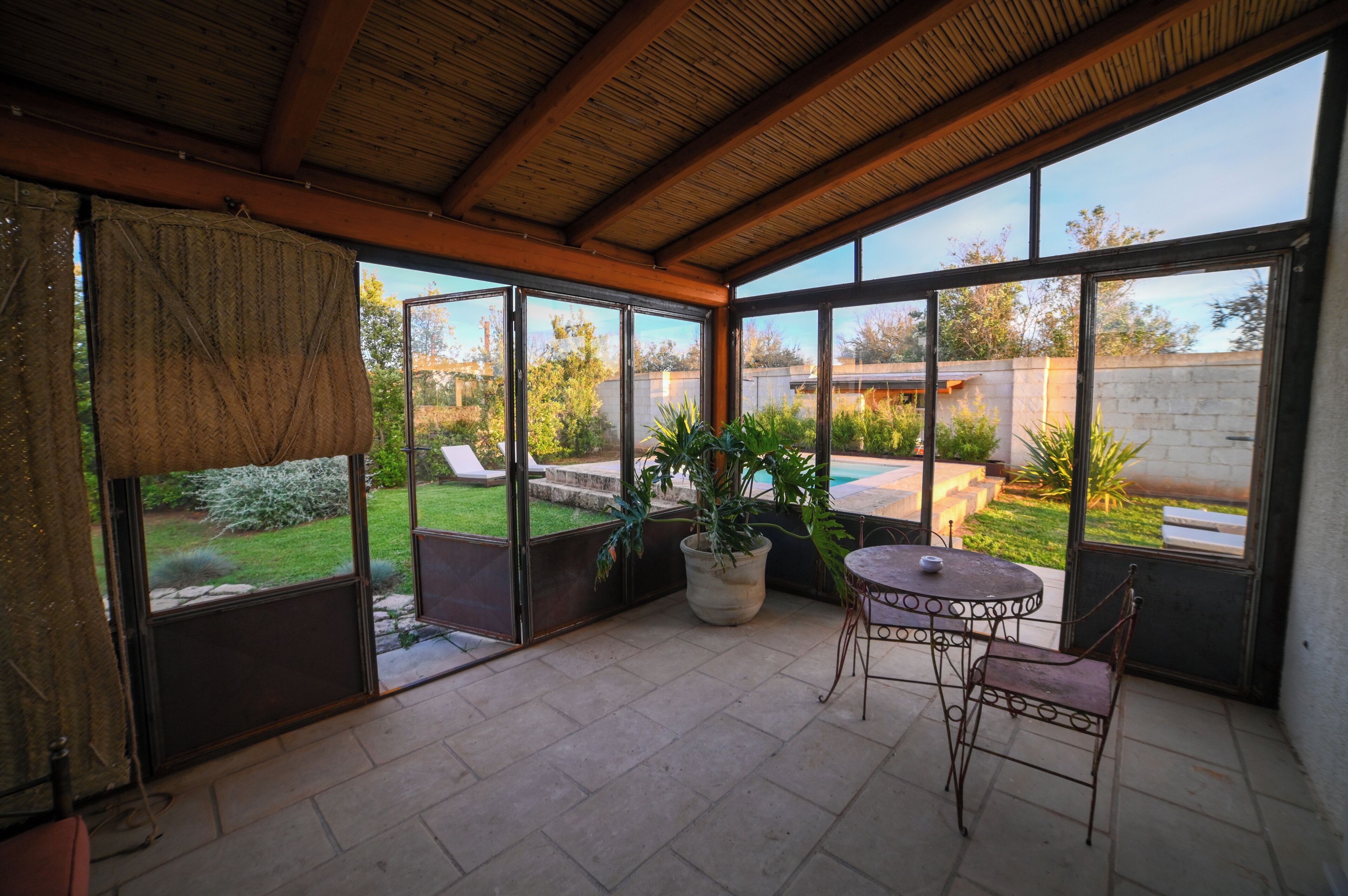 A covered patio with a table and chairs, large windows, and a view of a backyard with a pool and greenery.