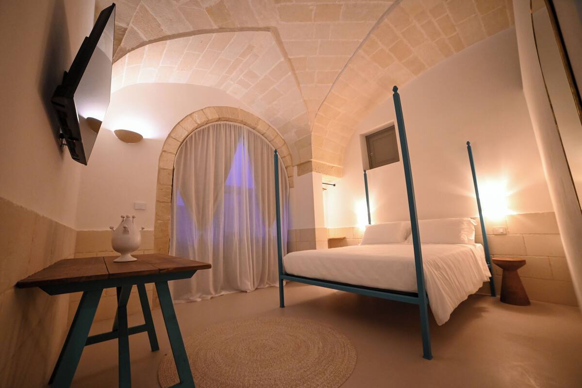 Une chambre à coucher avec un lit à baldaquin, un téléviseur à écran plat fixé au mur et une petite table surmontée d’un vase décoratif.