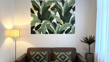 Una sala de estar con un sofá marrón, dos almohadas estampadas, una mesa de centro redonda de madera con una planta, una lámpara y un cuadro grande con hojas verdes.