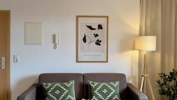 Una sala de estar con un sofá gris, dos cojines estampados, una pequeña mesa de centro de madera con un bonsái, una impresión botánica enmarcada en la pared, una lámpara de pie y una planta en maceta.