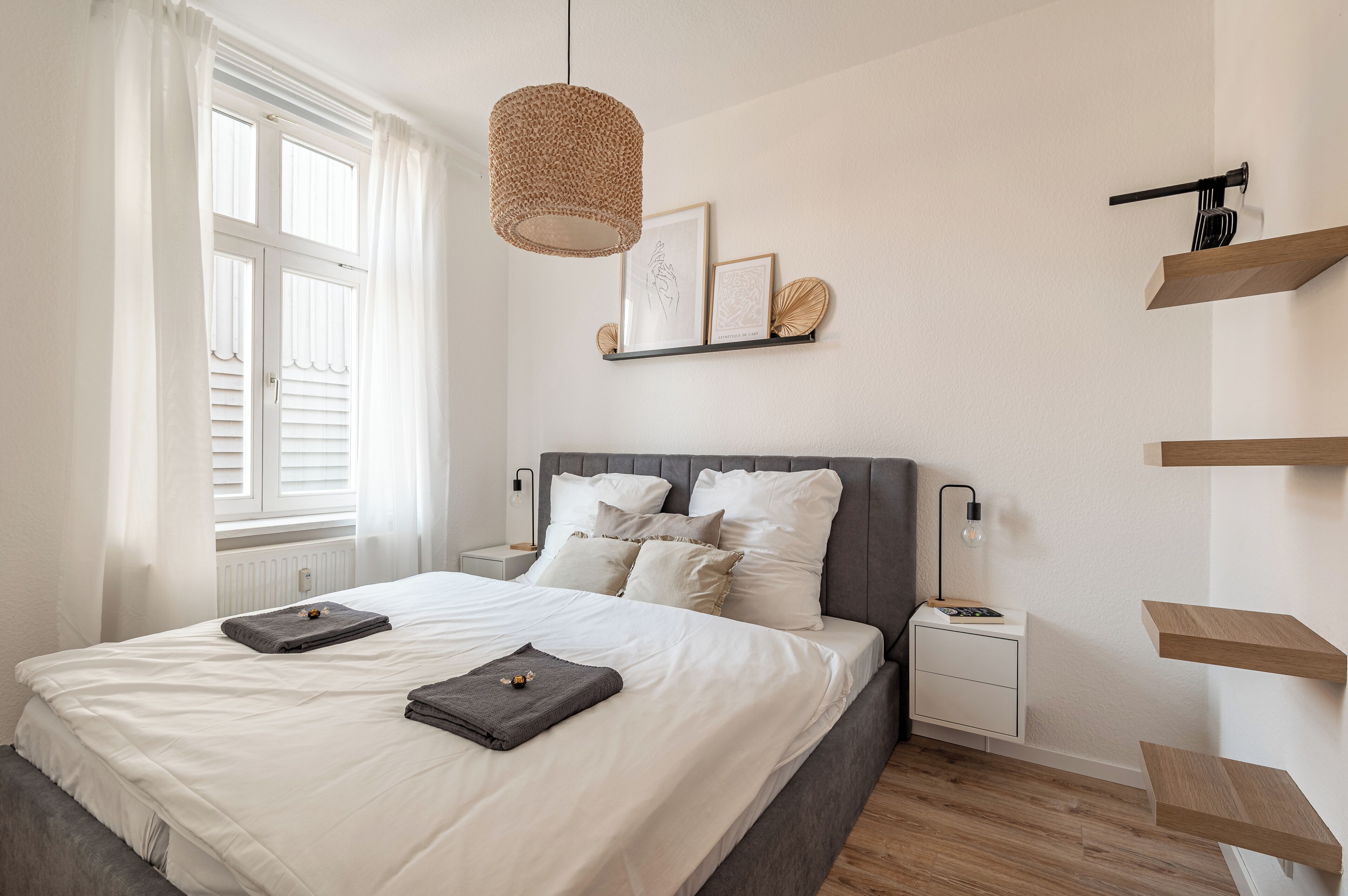 A bedroom with a bed, bedside tables, a hanging light fixture, and a wall-mounted shelf with decorative items.