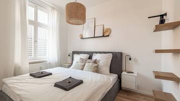 A bedroom with a bed, bedside tables, a hanging light fixture, and a wall-mounted shelf with decorative items.