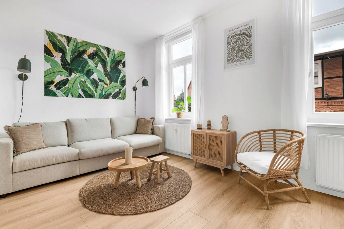 Una sala de estar con un sofá, una mesa redonda de madera, un sillón de mimbre e impresión botánica de gran tamaño en la pared.