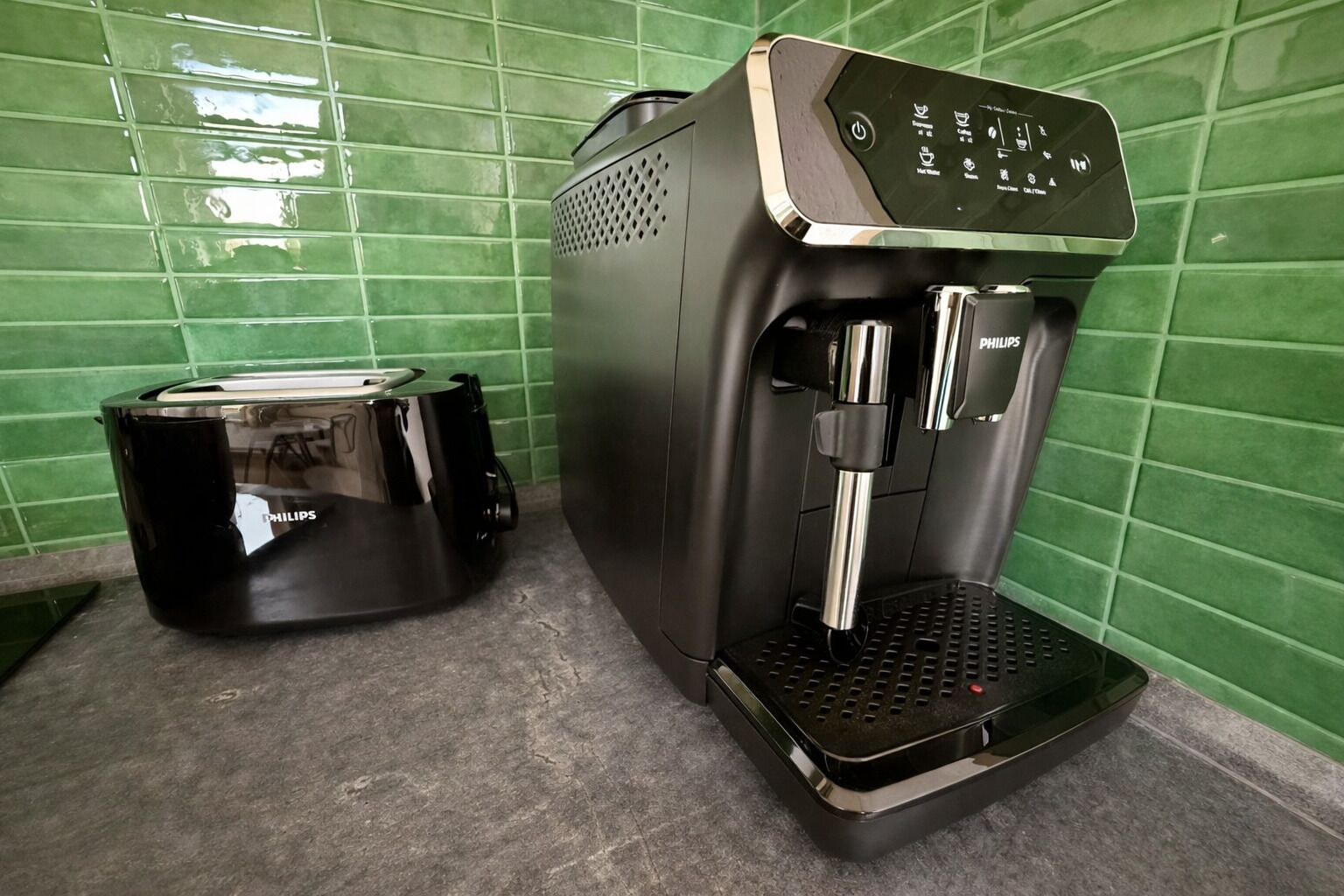 Eine Kaffeemaschine und ein Toaster auf einer Arbeitsplatte mit dunkelgrünen Fliesen.
