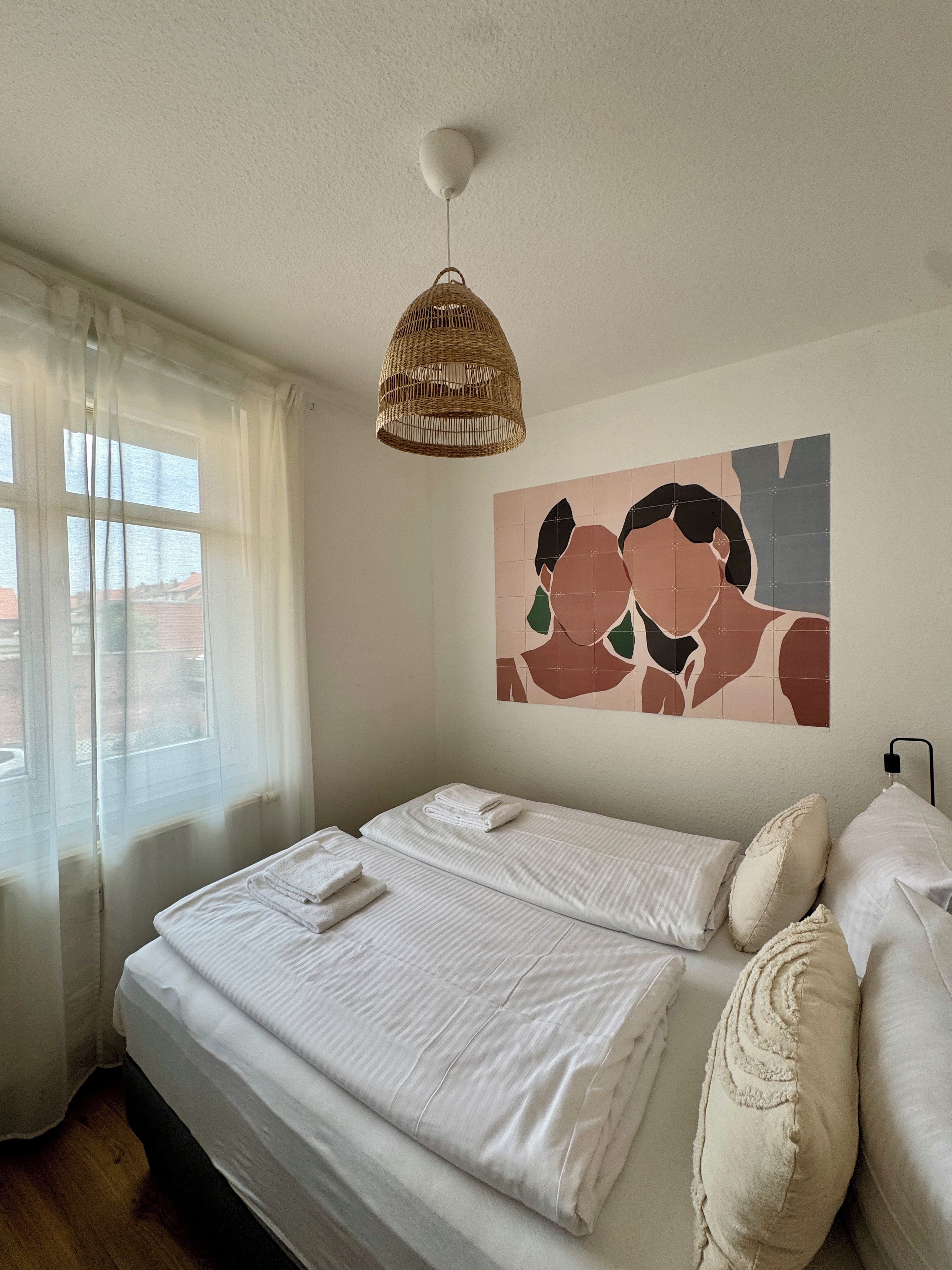 A neatly made bed with white linens and pillows, a woven pendant light, and a wall art piece.