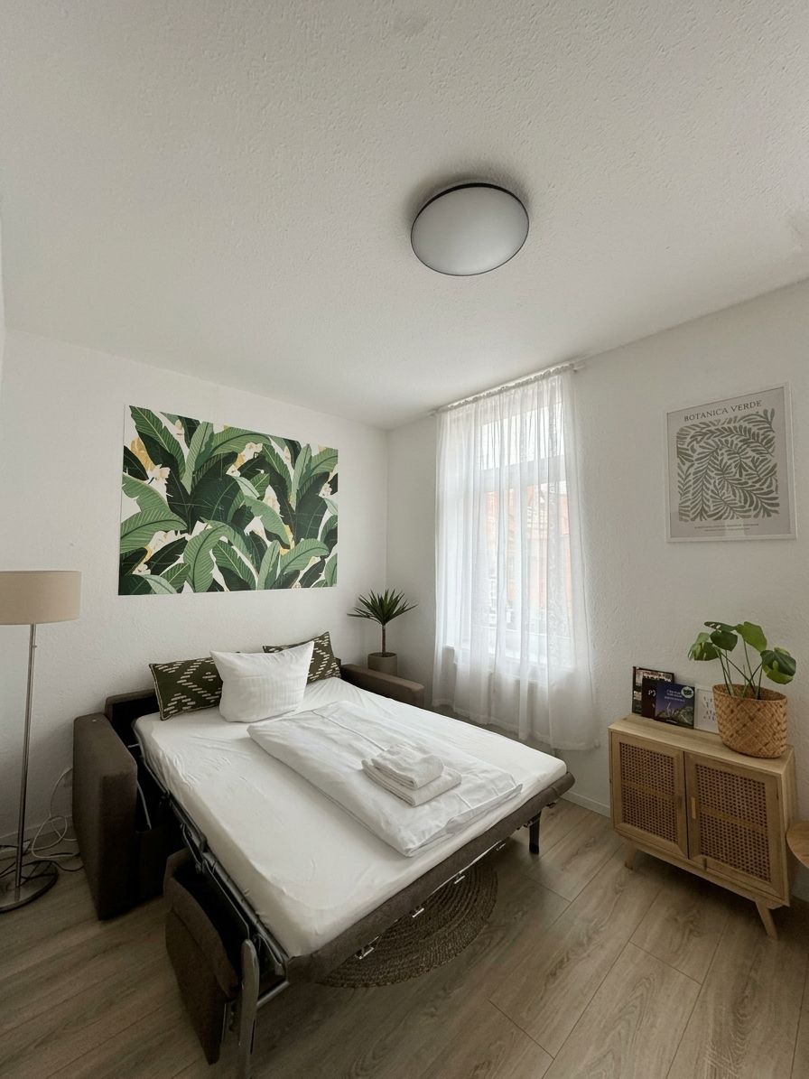 A hotel room with a bed, a bedside table, a lamp, a plant, and a window with curtains.