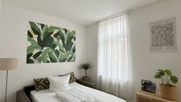 A hotel room with a bed, a bedside table, a lamp, a plant, and a window with curtains.