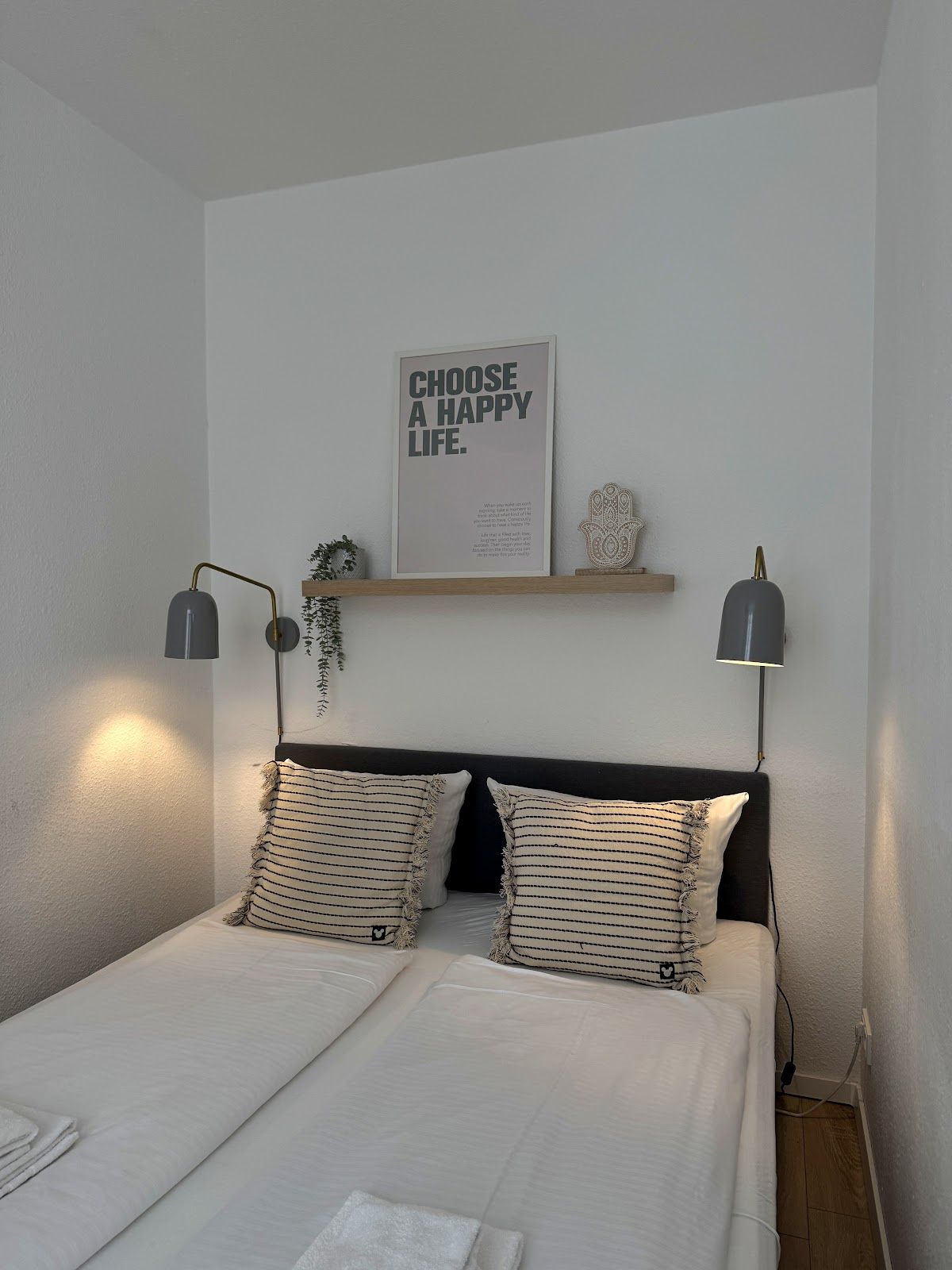 A bedroom with a bed, two bedside lamps, a wall-mounted shelf with decorative items, and a motivational poster.