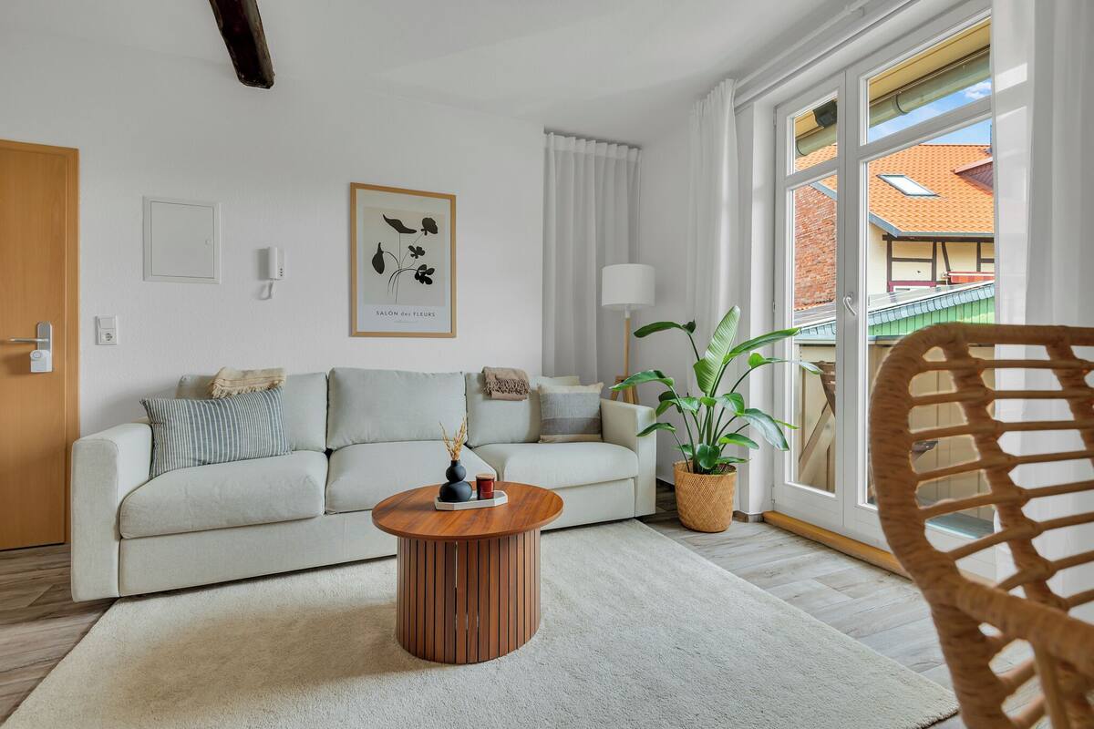 Una sala moderna con un sofá de color claro, una mesa de centro de madera, una planta en maceta y un amplio ventanal con vistas a un edificio.