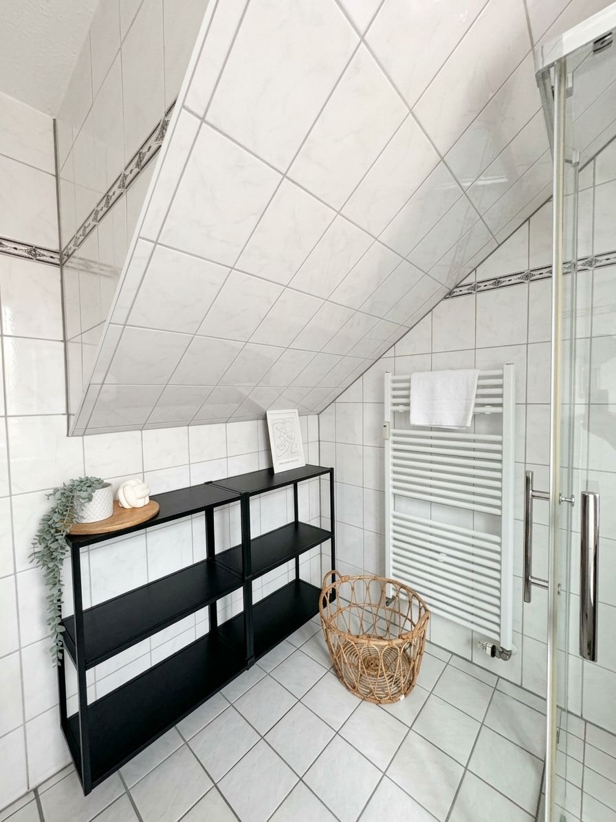 A bathroom with white tiled walls, a black shelf unit, a glass shower enclosure, and a white towel rack.