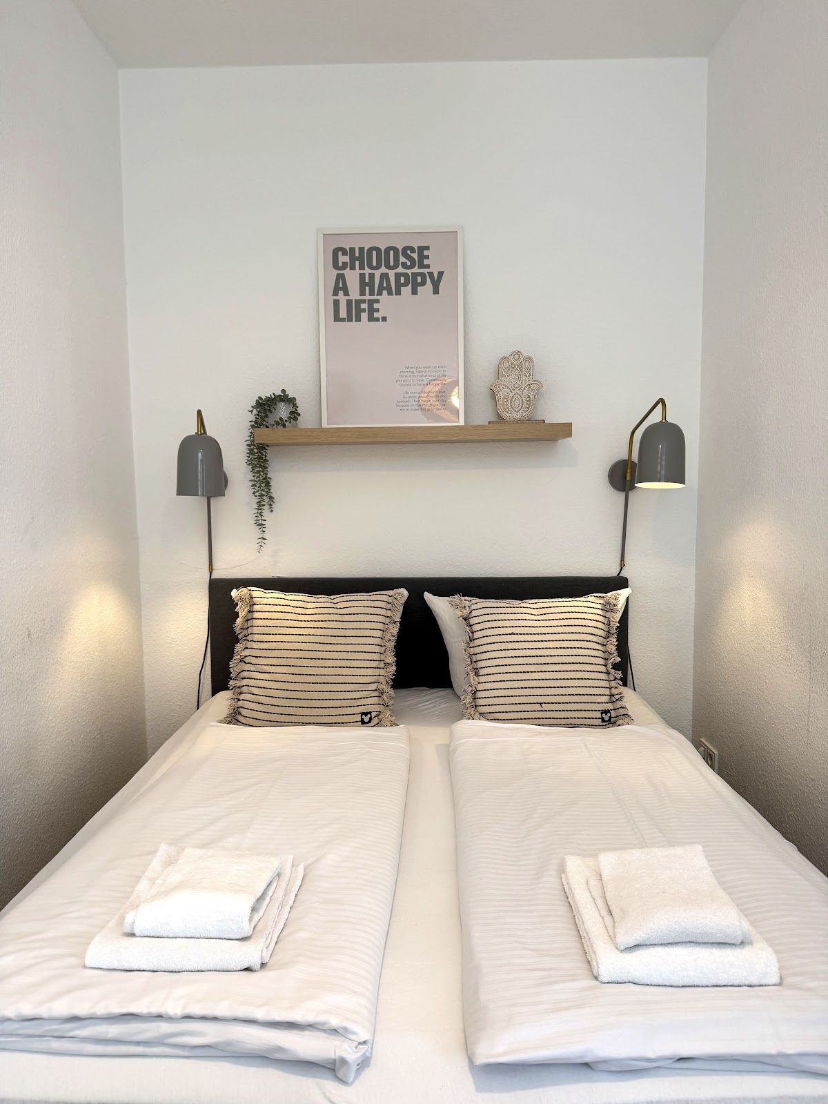 A double bed with two pillows, a bedside lamp, a wall-mounted shelf with a plant, and a motivational poster.