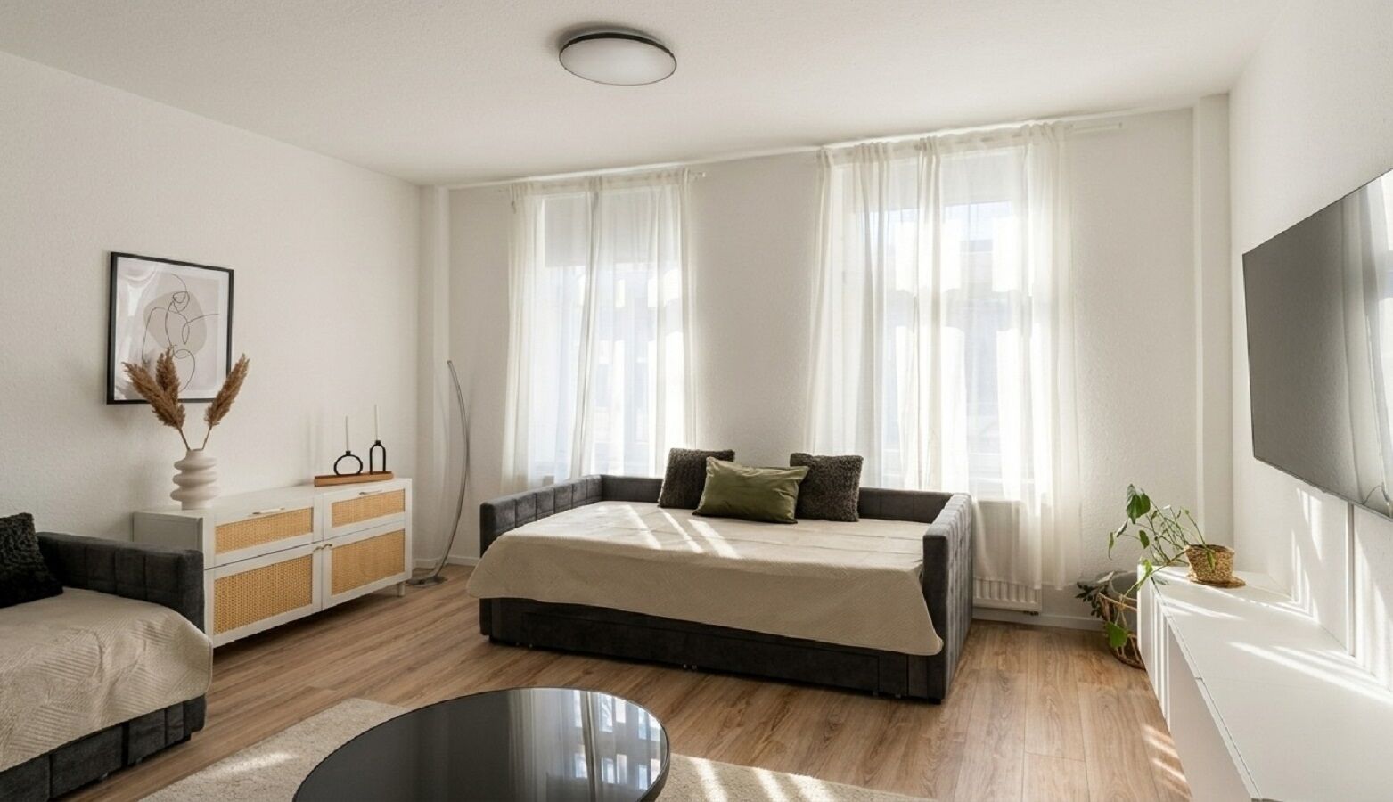 A modern bedroom with a large bed, a bedside table, a TV, and a window with sheer curtains.