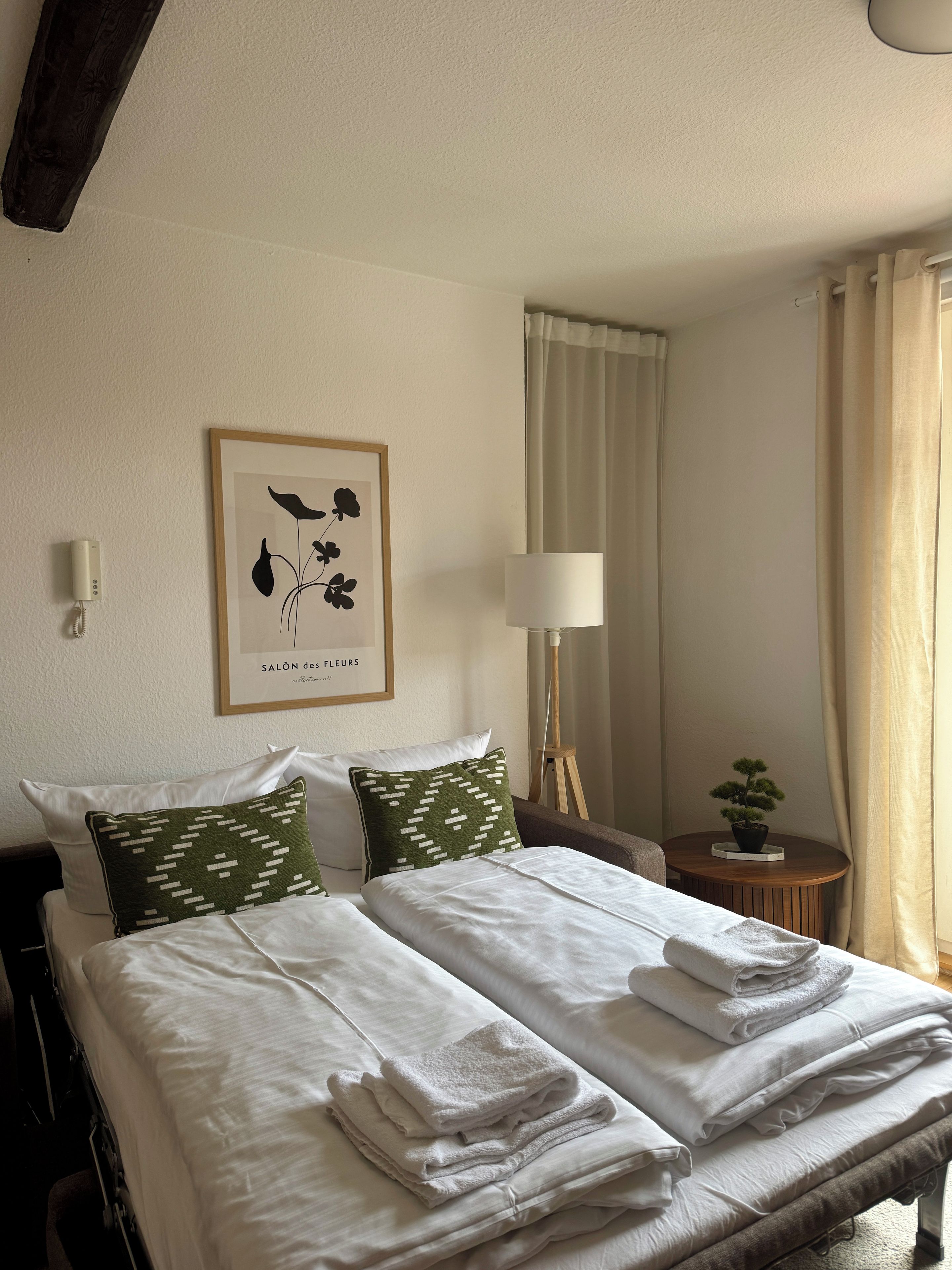Ein Hotelzimmer mit Bett, Kissen, einem gerahmten Kunstwerk, einer Nachttischlampe, einem kleinen Tisch mit Pflanze und einem Fenster mit Vorhängen.